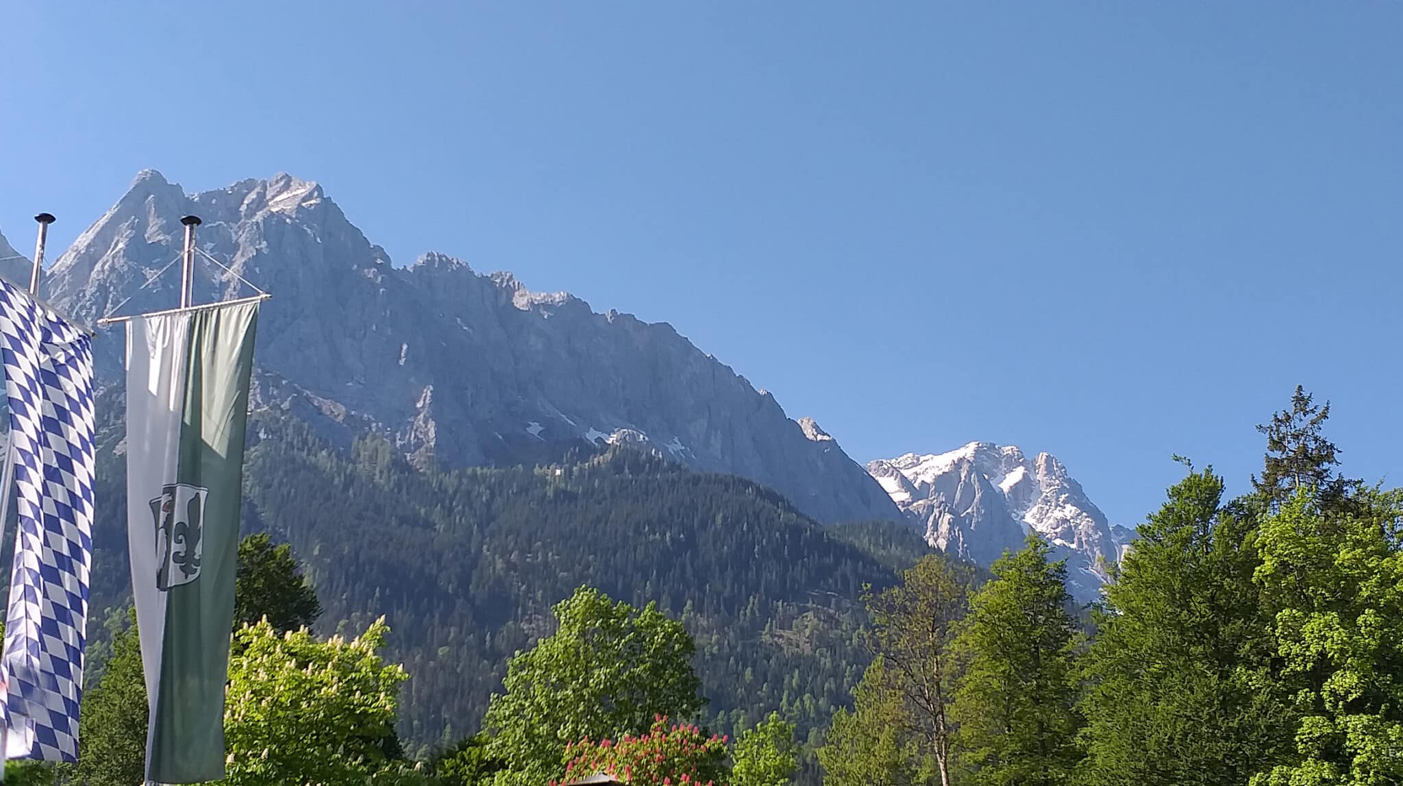 Im Hintergrund Großer Waxenstein und Zugspitze im Frühjahr. Im Vordergrund Fahnen von Bayern mit weiß-blauen Rauten und Grainauer Fahne.