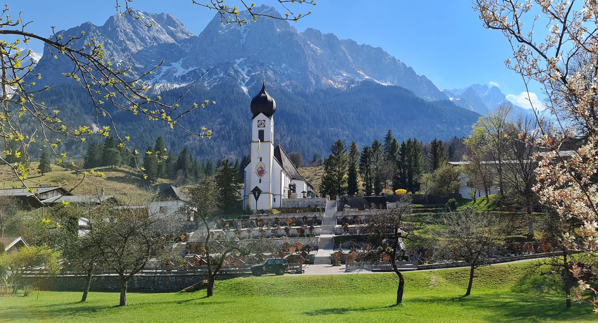 Grainauer Pfarrkirche mit Bergfriedhof im Frühjahr. Alpspitze, Waxensteine und Zugspitze schneebedeckt im Hintergrund