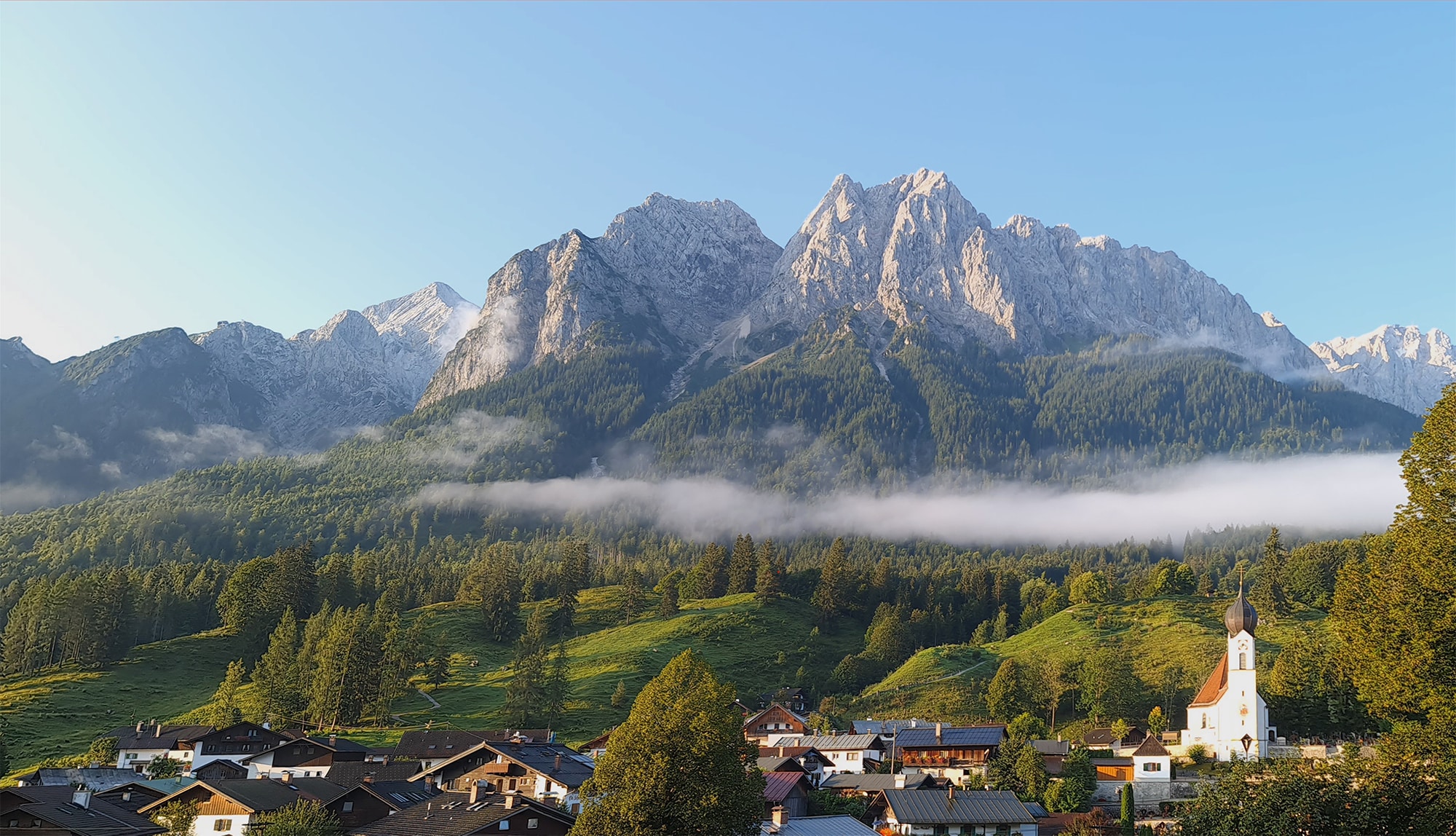 Grainau im Sommer. Blick vom Rathausbichl auf das Bergmassiv mit Alpspitze, Waxensteine und Zugspitze. Pfarrkirche und Dorf im Vordergrund