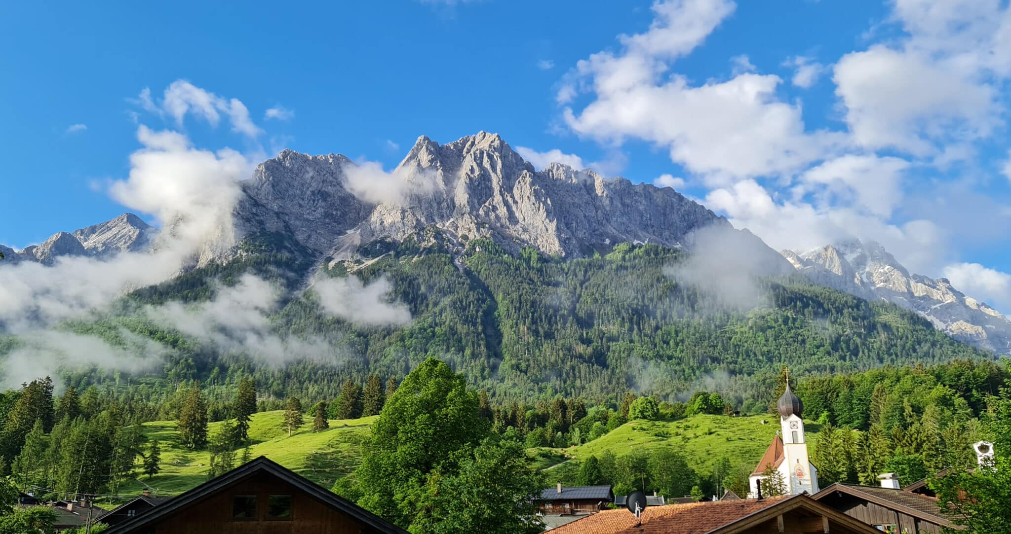 Grainau im Sommer. Blick vom Rathausbichl auf das Bergmassiv mit Alpspitze, Waxensteine und Zugspitze. Pfarrkirche und Dorf im Vordergrund