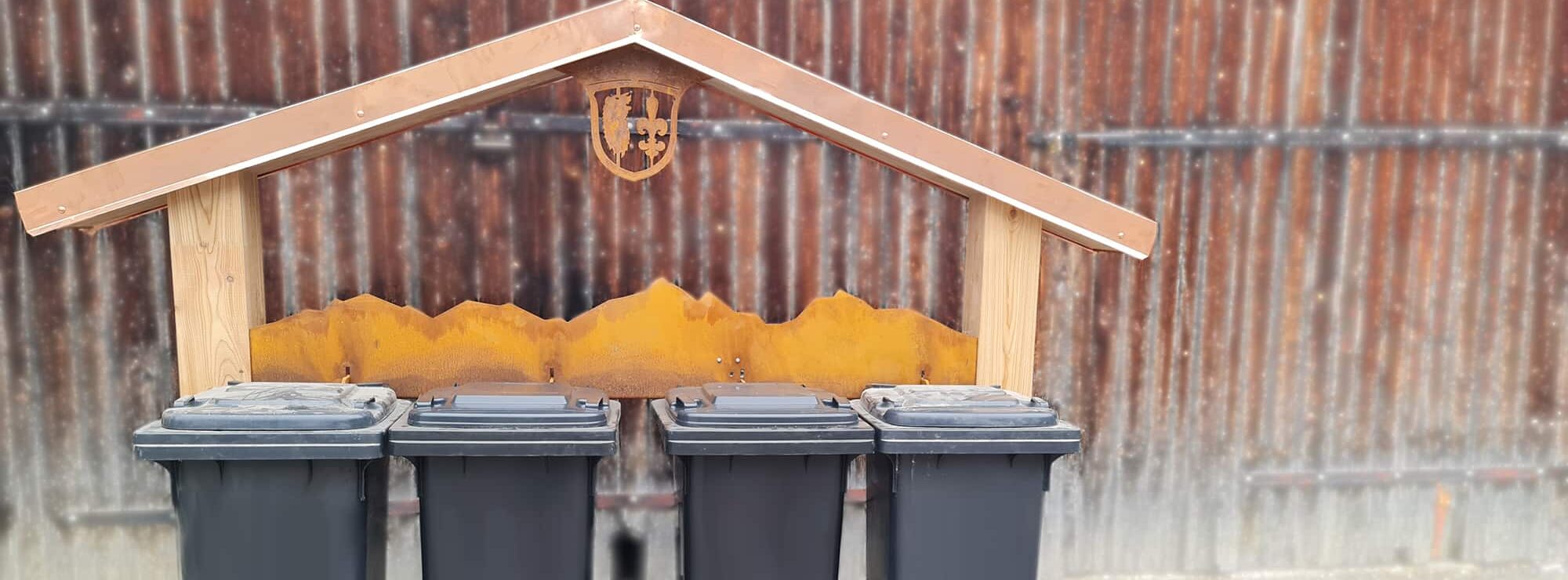 Grainau, Grainauer Müllstationen, vier Restmülltonnen im Vordergrund. Hintergrund Gestell aus Holz und Metall in Hausform mit Grainauer Wappen im Giebel, Stilisierte Berge aus Kupfer