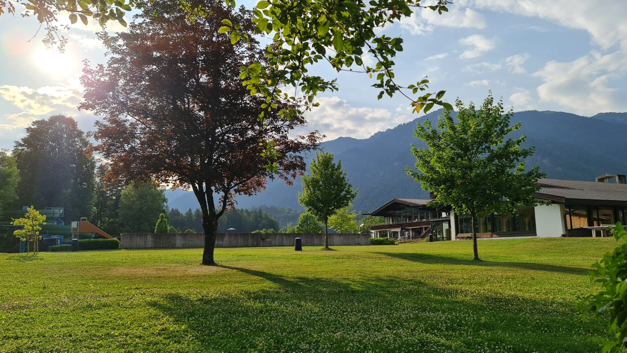 Grainau im Sommer. Das Zugspitzbad am Abend. Vordergrund Liegewiese mit Bäumen, Schwimmbadgebäude und Rutschen. Hintergrund Berge